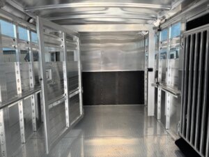 Interior of an empty Elite 30 metal livestock trailer with aluminum walls and floor, open gates, and a partition separating sections; sunlight streams through small windows—ideal for show cattle or as a versatile cattle combo setup. -Stillwater Trailer Sales