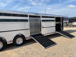 A 30' Elite Combo silver livestock trailer with two open side doors, each with a black ramp extended to the ground, is parked on gravel under a partly cloudy sky. The trailer is ideal for show cattle, featuring barred windows and a metal gate inside. -Stillwater Trailer Sales