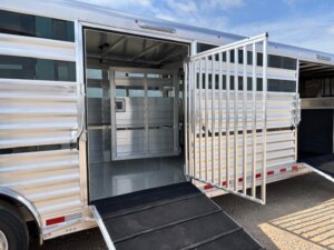 A silver Elite 30' Combo cattle equipment trailer with open side and interior gates, ramps extended to the ground, and a clean, empty interior, is parked on a dirt surface under a partly cloudy sky—ideal for hauling show cattle. -Stillwater Trailer Sales