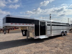A large silver and black Elite 30 livestock trailer with multiple axles is parked on a gravel lot under a partly cloudy sky. Featuring side vents and a gooseneck hitch, it’s ideal for transporting show cattle or as a versatile cattle combo. -Stillwater Trailer Sales
