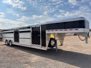 An Elite 30 Cattle Combo livestock trailer with three axles is parked on a gravel lot under a partly cloudy sky. This large, black and silver trailer features multiple doors and ventilation slats along the sides, ideal for show cattle transport. -Stillwater Trailer Sales