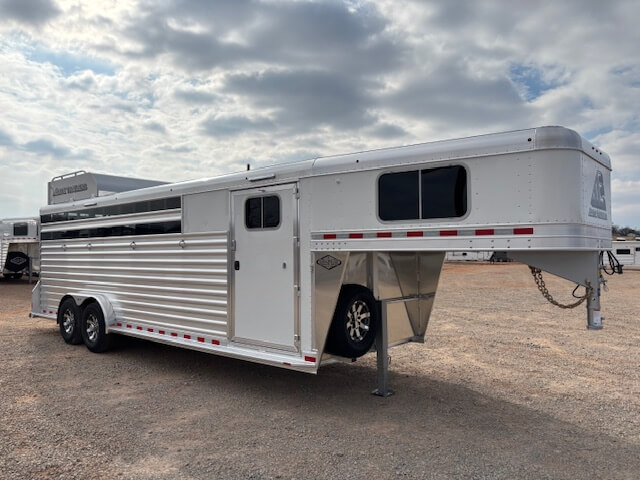 An Elite 24' Stock Combo by Elite Trailers, this silver gooseneck stock trailer features ventilation slats, a side door, and tandem axles, parked on a gravel lot under a partly cloudy sky. -Stillwater Trailer Sales