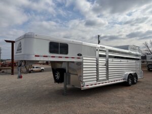 A large silver Elite 24' Stock Combo with a gooseneck hitch is parked on gravel under a cloudy sky. This livestock trailer features side vents, two axles, and a rear ladder. Other vehicles are visible in the background. -Stillwater Trailer Sales
