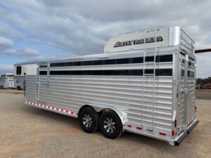 A large silver Elite 24' Stock Combo livestock trailer with two axles is parked on gravel under a cloudy sky. The trailer features horizontal slats, black-tinted windows, a ladder, and “Elite Trailers” branding on top. -Stillwater Trailer Sales
