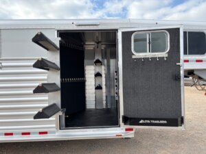 The image shows the open tack room of an Elite 24' Stock Combo trailer with saddle racks on the door and inside wall, bridle hooks on the far wall, and a rubber mat floor. The silver exterior has a window and Elite Trailers logo visible. -Stillwater Trailer Sales