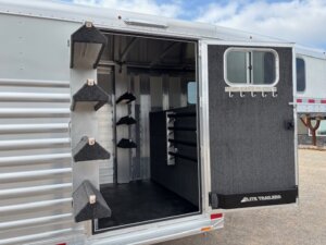 The image shows the open side door of an Elite 24' Stock Combo trailer, revealing an organized tack room with multiple saddle racks on the wall and door, bridle hooks, and carpeted interior walls. The stock trailer’s exterior is metallic with horizontal panels. -Stillwater Trailer Sales