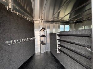 Interior of an Elite 24' Stock Combo horse trailer tack room with lined walls, multiple hooks on the left, three saddle racks at the far end, and several padded vertical racks on the right. A small window is visible on the right side. -Stillwater Trailer Sales