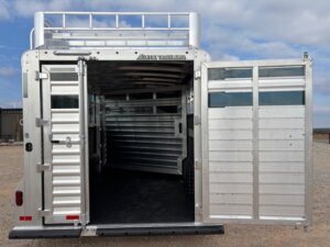 An open silver Elite 24' Stock Combo livestock trailer with one rear door fully open and the other partly open, revealing an empty interior, set on a gravel surface under a partly cloudy sky. -Stillwater Trailer Sales
