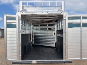 A silver Elite 24' Stock Combo livestock trailer with open rear double doors reveals an empty, clean interior featuring black rubber flooring and metallic walls. The trailer is parked outdoors under a partly cloudy sky. -Stillwater Trailer Sales