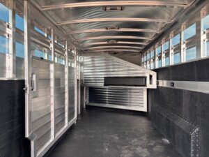 Interior of an empty Elite 24' Stock Combo trailer with aluminum walls, roof, and gates. The stock trailer features small side windows for ventilation and a non-slip black floor, with light entering from the sides and rear. -Stillwater Trailer Sales
