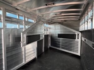 The interior of an empty Elite 24' Stock Combo livestock trailer features ribbed metal walls, a black rubber floor, overhead structural beams, and two black feed troughs on angled partitions. Natural light enters through side windows. -Stillwater Trailer Sales