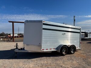 A silver, enclosed CM 16' Bumper Pull livestock trailer with two axles is parked on gravel near a road. The trailer features horizontal ridges, a black and white stripe, and a small logo on its side. The sky is mostly clear with few clouds. -Stillwater Trailer Sales