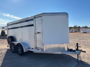 A silver, enclosed CM 16' Bumper Pull livestock trailer with dual axles is parked on a gravel lot. The trailer features a vented side, front hitch, and sits under a clear blue sky with other trailers visible in the background. -Stillwater Trailer Sales