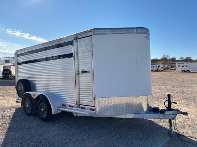 A silver CM 16' Bumper Pull livestock trailer with two axles and black wheels is parked on a gravel lot. The trailer has a vented upper section, side door, and hitch for towing. Several other trailers are visible in the background. -Stillwater Trailer Sales