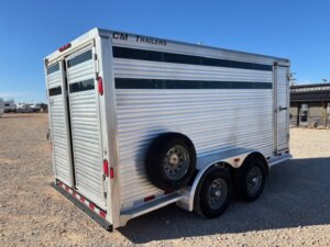 A silver CM 16' Bumper Pull livestock trailer with two axles and a spare tire mounted on the side is parked on gravel in an open lot. The trailer features a corrugated metal exterior and rear doors with vents. -Stillwater Trailer Sales