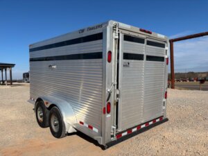 A silver CM 16' Bumper Pull livestock trailer with two axles is parked on a gravel lot under a clear blue sky. The trailer features horizontal ventilation slats and rear double doors. -Stillwater Trailer Sales