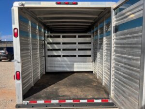 The image shows the interior of an empty CM 16' Bumper Pull aluminum livestock trailer with ribbed walls, a non-slip floor, and a central divider gate. The trailer is parked outdoors on a sunny day. -Stillwater Trailer Sales