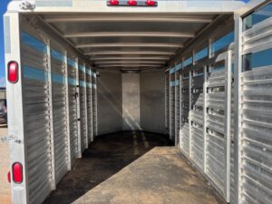 Interior view of an empty CM 16' Bumper Pull trailer with ribbed metal walls, open side slats for ventilation, and a non-slip floor. The well-lit CM trailer has open rear doors for easy access. -Stillwater Trailer Sales