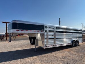 An Elite 24' show cattle trailer with a gooseneck hitch is parked on gravel. The large, silver cattle trailer features slatted sides, tandem axles, and sits beneath a clear blue sky. -Stillwater Trailer Sales