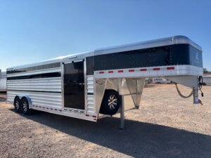A silver and black Elite Cattle Trailer with tandem wheels is parked on gravel under a clear blue sky. The 24' cattle trailer features side slats, an open side door, and a spare tire mounted inside the front section. -Stillwater Trailer Sales