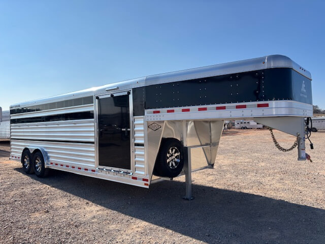 An Elite 24 Cattle Trailer in silver and black with double axles is parked on gravel under a clear blue sky. The gooseneck livestock trailer features side vents, a door, and a spare tire mounted inside the gooseneck area. -Stillwater Trailer Sales
