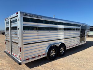 A large, silver Elite 24' Cattle Trailer with black and chrome wheels is parked on a gravel lot under a clear blue sky. The trailer features side vents and is labeled “Elite Trailers” at the top. -Stillwater Trailer Sales