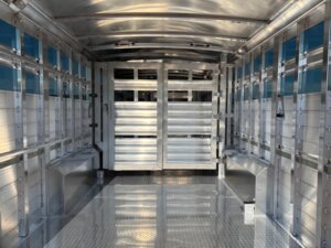 Interior view of an empty Elite 24' Show Cattle Trailer with reflective metallic walls, ventilated side panels, a ridged floor, and closed double doors at the far end. -Stillwater Trailer Sales