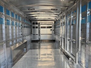 Interior view of an empty Elite 24' Show Cattle Trailer with a diamond plate floor, multiple slatted windows along both sides, and a closed gate at the far end. -Stillwater Trailer Sales