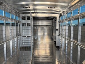 Interior view of an empty Elite 24' cattle trailer with barred sides, open rear doors, a small side door, and diamond-plate flooring. The well-lit Show Cattle Trailer is illuminated by natural light streaming in from outside. -Stillwater Trailer Sales
