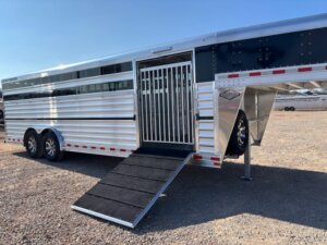 A silver and black Elite 24' show cattle trailer with two axles, an open side door with vertical metal bars, and a black ramp extended to the ground, is parked on a gravel lot under a clear sky. -Stillwater Trailer Sales