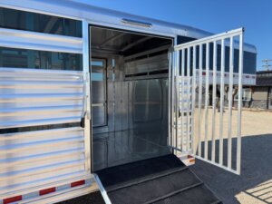 A silver 24' cattle trailer with its rear gate and side door open, showing a metal ramp leading inside. The interior is empty and clean, with sunlight reflecting off the polished surfaces. -Stillwater Trailer Sales
