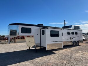 A large white Trails West 3 Horse LQ horse trailer with multiple windows, a slide-out section, and shiny metal trim is parked on gravel under a clear blue sky. -Stillwater Trailer Sales