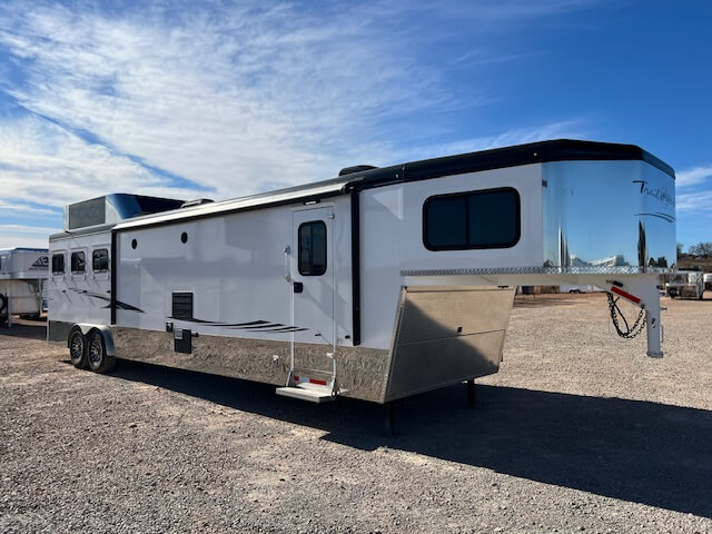 A large white and silver Trails West gooseneck horse trailer with multiple windows, black trim, and dual axles is parked on a gravel lot under a blue sky with scattered clouds. -Stillwater Trailer Sales