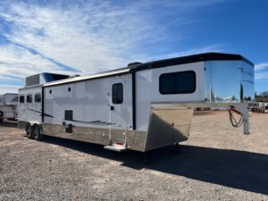 A large white Trails West fifth-wheel horse trailer with black accents and two axles is parked on a gravel lot under a blue sky with scattered clouds. The trailer has tinted windows and a side door. -Stillwater Trailer Sales