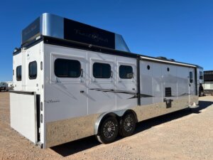 A large, white Trails West horse trailer with multiple windows and black detailing is parked on a gravel lot under a clear blue sky. This 3 Horse LQ trailer features dual axles, chrome wheels, and a prominent storage compartment on top. -Stillwater Trailer Sales