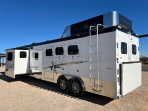 A large white and silver Trails West travel trailer with multiple windows, a side awning, a rear ladder, and shiny chrome wheels is parked on a gravel surface under a clear blue sky. -Stillwater Trailer Sales
