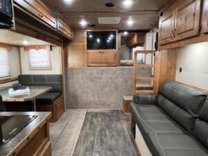 Interior of a Trails West 3 Horse LQ horse trailer with wood cabinetry, a gray sofa on the right, small dining benches on the left, TV above storage, raised bed area in back, and vinyl wood-look flooring. -Stillwater Trailer Sales