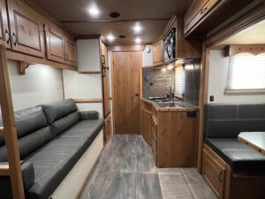 The interior of this Trails West 3 Horse LQ horse trailer features a gray couch on the left, a small kitchen with a sink and microwave in the center, a wooden door at the back, and a dining booth with gray cushions on the right. -Stillwater Trailer Sales