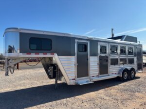 A large, silver Cimarron gooseneck 4 Horse Trainer trailer with four side windows, multiple doors, and tandem axles is parked on a gravel lot under a clear blue sky. -Stillwater Trailer Sales
