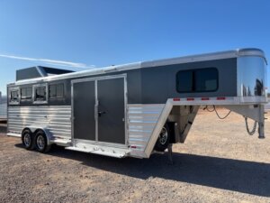 A silver and gray Cimarron gooseneck horse trailer with three windows and dual axles is parked on gravel under a clear blue sky. Perfect for a 4 Horse Trainer’s needs. -Stillwater Trailer Sales