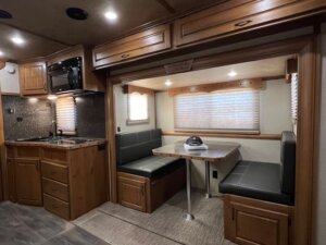 An RV interior inside a Trails West horse trailer features wood cabinetry, a kitchenette with microwave and sink on the left, and a dinette on the right with two bench seats, table, windows with blinds, and a hat resting on the table. -Stillwater Trailer Sales