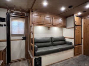 Interior of a Trails West 3 Horse LQ Horse Trailer featuring a gray sofa against a wall with overhead wooden cabinets, a window with a closed blind, wood-paneled refrigerator, storage compartments, and door. -Stillwater Trailer Sales