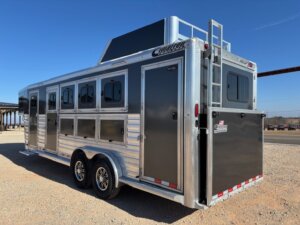 A shiny silver and black Cimarron 4 Horse Trainer gooseneck trailer with multiple windows and doors is parked on a gravel lot under a clear blue sky. A ladder is attached at the rear, and it has two axles with alloy wheels. -Stillwater Trailer Sales