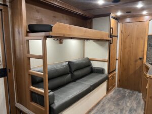 A compact Trails West LQ RV interior features a wooden bunk bed above a gray cushioned sofa, wood-paneled walls and door, overhead lights, and laminate flooring. The area appears clean and well-maintained—perfect for 3 Horse adventures. -Stillwater Trailer Sales