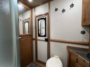 A small bathroom in a Trails West 3 Horse LQ horse trailer features wood-paneled walls, a white toilet, a shower with frosted door, black countertop and sink, mirrored door, and black wall hooks. -Stillwater Trailer Sales