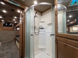 A compact shower with frosted sliding glass doors, a handheld showerhead, and built-in shelves is shown inside a Trails West 3 Horse LQ horse trailer bathroom. The space features wood cabinets, gray flooring, and recessed ceiling lights. -Stillwater Trailer Sales