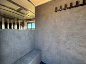 The image shows the interior of a Cimarron 4 Horse Trainer trailer with gray carpeted walls, a small bench seat, a window, and a black rack with hooks. The space appears clean and empty. -Stillwater Trailer Sales