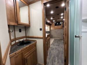 View of a Trails West 3 Horse LQ horse trailer interior, featuring a small wooden vanity with black sink on the left, wood cabinets, laminate flooring, and a doorway leading to a sleeping area with bed and mounted TV in the background. -Stillwater Trailer Sales