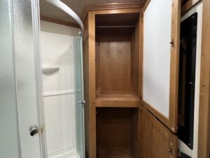 A wooden cabinet with an open door stands next to a glass shower enclosure in a Trails West 3 Horse LQ horse trailer bathroom. The cabinet has a shelf inside, is built into the wall, and features additional storage compartments nearby. -Stillwater Trailer Sales