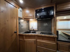 A compact kitchen in this 3 Horse LQ horse trailer features wooden cabinets, a black microwave, a double sink, a stove with a closed cover, and a window with blinds. The speckled countertop includes built-in drawers below. -Stillwater Trailer Sales